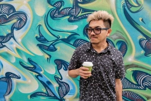 Asian man holding a latte standing in front of a green and blue picture